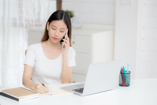 Young asian woman talking phone and work from home with laptop computer, freelance girl writing on notebook and speak on smartphone, stay home, domestic life, business and communication concept.