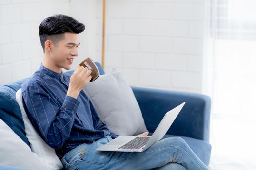 Young asian businessman smile and work from home with laptop computer online on sofa in living room, freelance business man using notebook and drink coffee on couch, new normal, lifestyle concept.