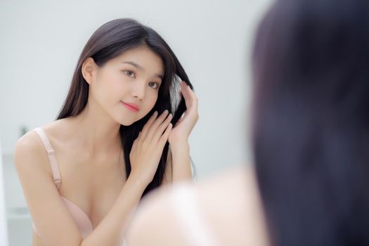 Beautiful young asian woman smiling look at mirror of touch hair in the room, cosmetic for hygiene hair care, beauty asia girl happy for hairstyle treatment at home, lifestyle and makeup concept.