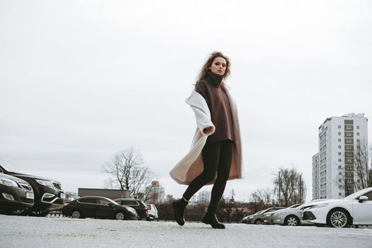 A girl with red curly hair in a white coat poses on outdoor parking in cold autumn. City Style - Urban