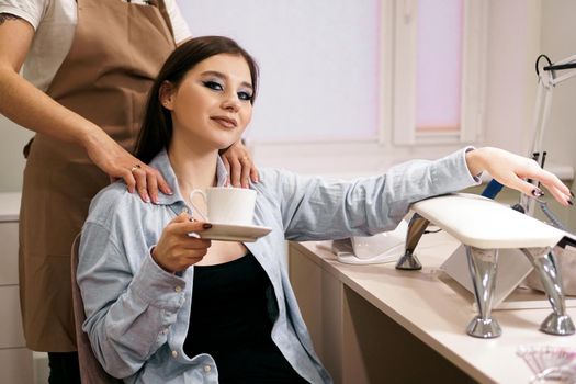 Beautiful woman getting manicure and massage at the same time in a beauty salon. She smiles and holds a cup of coffee. VIP client in a beauty salon