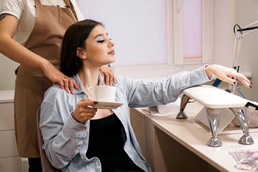 Beautiful woman getting manicure and massage at the same time in a beauty salon. She smiles and holds a cup of coffee. VIP client in a beauty salon