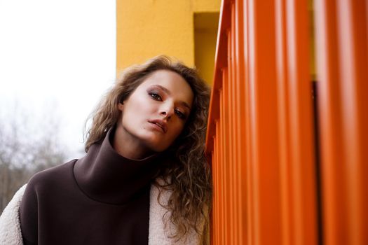 A girl with red curly hair in a white coat poses on the yellow orange parking stairs. City Style - Urban