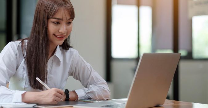 A young, cheerful girl in a white shirt smiles, smiling toothily writing down notes holding training for students to be executives at laptop desktop table