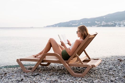 Summer vacation on the beach. The beautiful young woman in swimming suit sitting on the sun lounger reading a book and relaxing