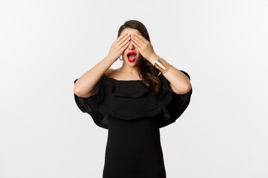 Fashion and beauty. Beautiful woman in black dress and red lipstick, covering eyes and smiling, waiting for surprise, standing over white background.