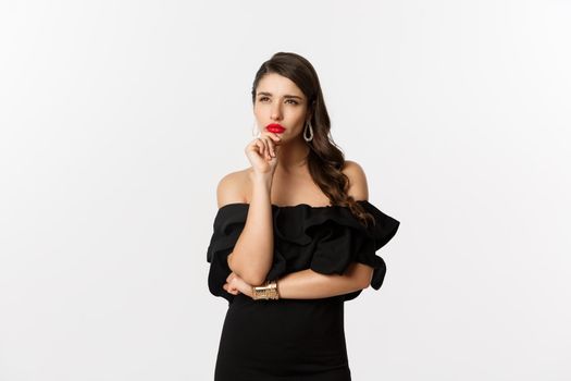 Fashion and beauty. Image of thoughtful glamour woman looking away, squinting while thinking, standing in black dress against white background.