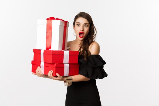 Celebration and christmas holidays concept. Beautiful woman in black dress holding gifts and looking surprised, standing over white background.