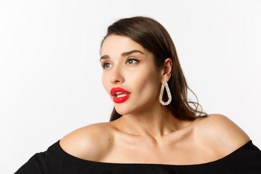 Fashion and beauty concept. Close-up of luxury woman with red lips, earrings and black dress, looking away sensual, standing over white background.