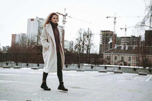 A girl with red curly hair in a white coat poses on outdoor parking in cold autumn. City Style - Urban. City construction in the background