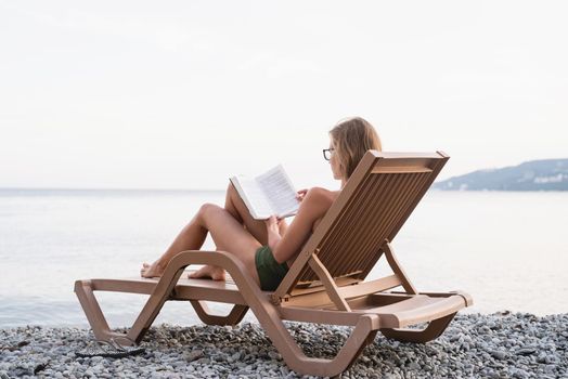 Summer vacation on the beach. The beautiful young woman in swimming suit sitting on the sun lounger reading a book and relaxing