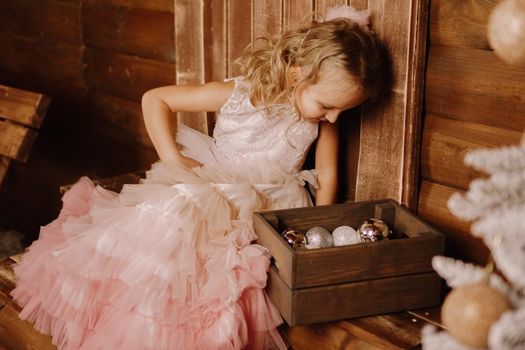 Cute girl with wavy hair in a pink elegant dress looking at a wooden box with christmas balls near the christmas tree