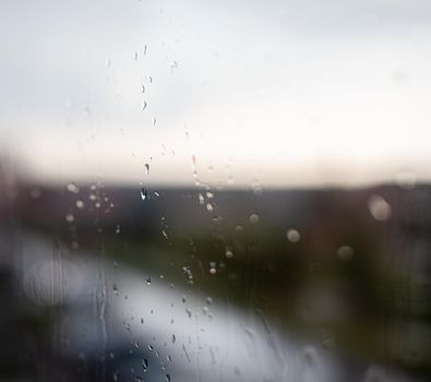 Rainy day through the window on cloudy grey sky and city buildings background. Concept. Evening cityscape behind the glass window with trickling drops of water.