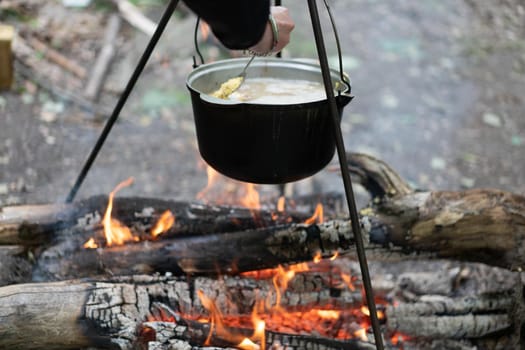 Camping outdoors. Cooking bowler hat hung on tripod over burning fire on the background of grass and chipped firewood