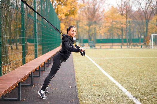 Girl athlete training using sport ground Mixed race young adult woman workout suspension system Healthy lifestyle Stretching outdoors playground. Make your body machine