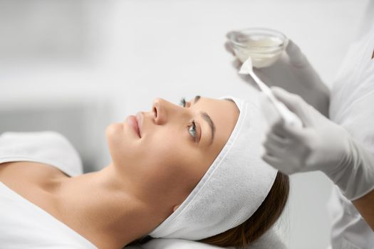 Side view of young beautiful brunette woman lying and beautician in rubber gloves doing cleaning face. Concept of special beauty procedure for improvements skin with different mask for face. 