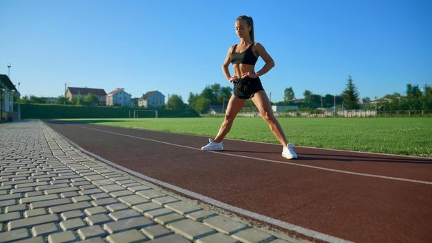 Side view of young sexy woman with perfect muscular body stretching before training at stadium in summer sunny morning. Gorgeous fit girl posing with legs wide and hands on waist outdoors.