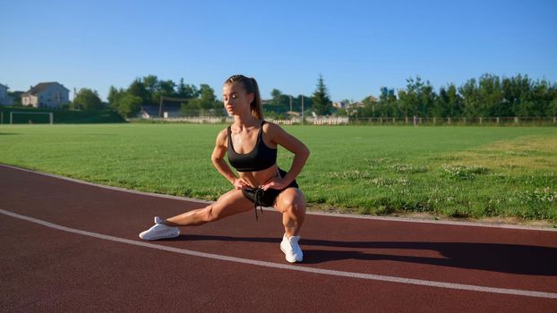 Fit flexible girl stretching legs, rolling from side to side outdoors. Sexy, muscular young woman warming up at stadium, practicing side lunges in sunny summer morning. Sport concept.