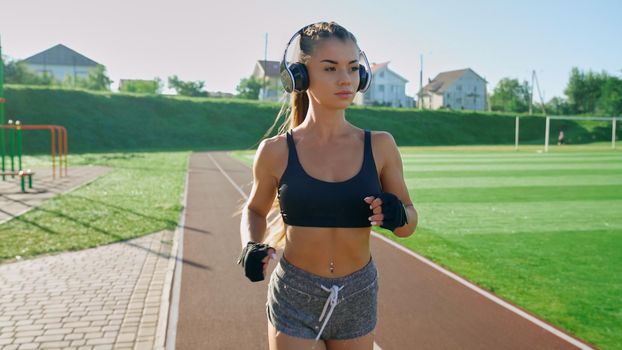 Front view of young athlete woman wearing big wireless headphones and black gloves running outdoors. Stunning girl with pony tail practicing jogging at stadium in sunny summer morning.