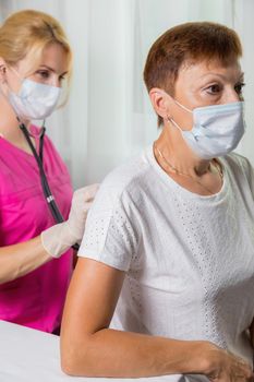 A girl doctor in a mask listens with a stethoscope to the back of an adult woman. A woman in the doctor's office complains of a cough