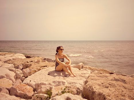 Beautiful young girl sitting on the rocks at the beach near the mediterranean sea. Summer season concept and barefoot caucasian woman relaxing and stay thoughtful on the coast