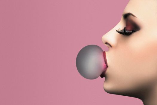 Beautiful woman with perfect makeup blowing pink bubble gum. Closeup portrait on a pink background with copy space