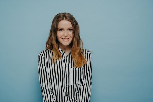 Lovely young female dressed casually with long hair looking positively at camera, studio shot of cute happy teenage girl in striped shirt expressing positive emotions while posing on blue background