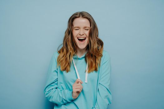 Portrait of optimistic teenage girl in casual clothes with tongue piercing keeping eyes closed and laughing joyfully, expressing happiness while posing isolated over blue background in studio