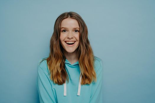 Studio shot of attractive happy young teenage girl in casual clothes looking at camera with happy face expression, showing positiveness, optimistic female wearing hoodie looking at camera and laughing