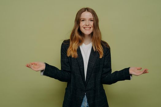 Smiling positive female shrugging shoulders with spreaded hands and looking at camera with positive face expression, does not know what to choose while posing against green studio background