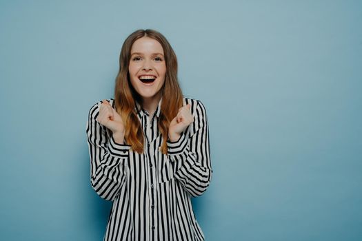 Overjoyed young woman wearing casual clothes feeling very happy and excited, doing winner gesture with clenched, screaming while celebrating success while posing isolated on blue background