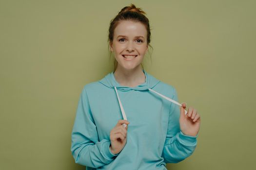 Portrait of cute flirty teenage girl biting lower lip playing with hoodie strings, standing in front camera on color background in studio, female feeling playful and carefree