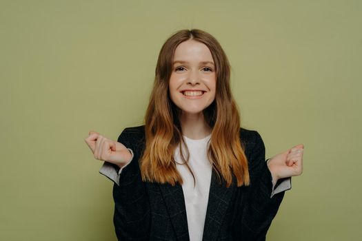Excited teenage girl with wavy hair in casual clothes celebrating victory and success, being very happy, smiling teenager doing winner gesture while standing isolated over green background