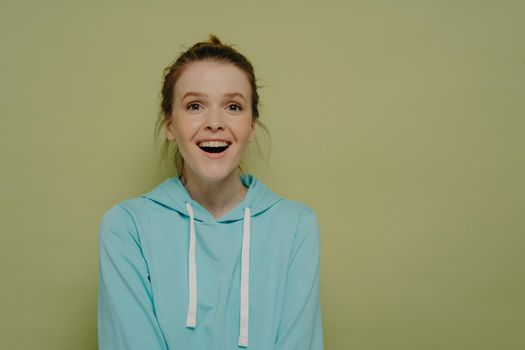 Amazed teenage girl with hair up wearing turquoise hoodie being excited after seeing something amazing, wow face expression and open mouth from amazement. Human emotions concept