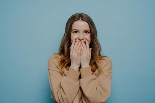 Can not believe my eyes. Amazed european teenage girl in casual clothes laughing and covering mouth with hands expressing positive emotions while posing against blue wall in studio