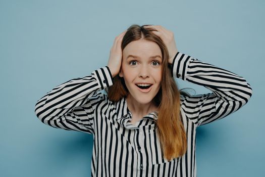 Surprised happy teenage girl standing in studio, holding head in hands and looking at camera with surprised face expression and opened mouth, female being excited when hearing about big sale prices