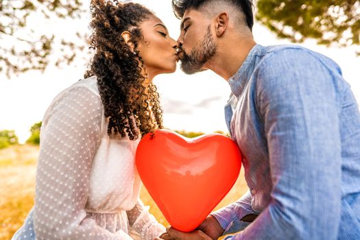 Profile photograph of multiracial couple in love kissing at sunset in nature with sun backlit effect on red heart shaped balloon among them. Romantic scene at dusk of two heterosexual young people