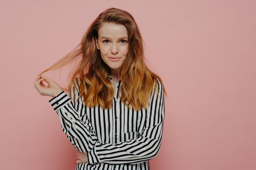 Beautiful young caucasian woman dressed in casual clothes playing with hair and looking at camera, cute female without makeup in shirt posing isolated on pink background. Studio shot