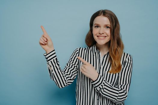 Cute teenage girl in striped shirt with shining smile pointing up with forefingers at copy space for advertisement, positive female advertising product while standing isolated on blue background
