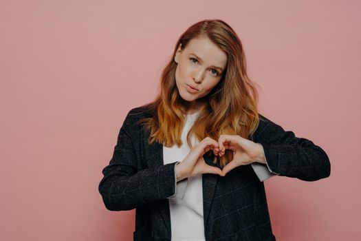 Beautiful young female with wavy ginger hair in formal dark jacket and white top showing heart shape sign tilting head and posing against light pink studio background. Body language concept