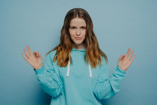 Confident smiling young girl with wavy brown hair showing okay sign with both hands while posing in comfortable blue sweater on blue studio background. Body language concept