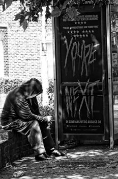 Homeless man sitting next to a telephone kiosk advertising a film. London, UK.