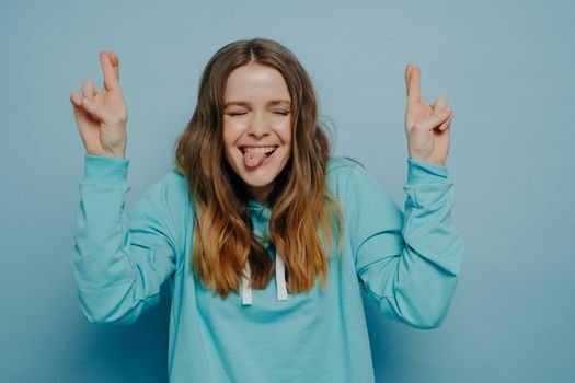 Happy crazy teenage girl with wavy ombre hair keeping fingers crossed while closing eyes and sticking tongue out standing against blue wall in studio, having fun and feeling carefree