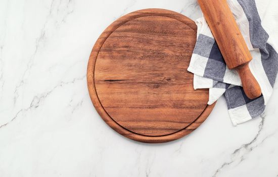 Empty wooden pizza platter with napkin and rolling pin set up on marble stone kitchen table. Pizza board and tablecloth on white marble background.