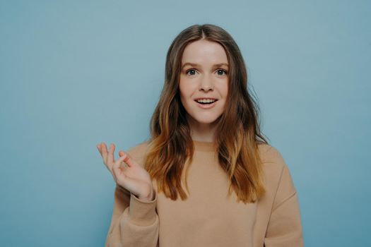 Smiling teenage girl raising hand and looking ahead with amusement and surprise, brunette with wavy hair wearing casual light brown sweater posing against blue background