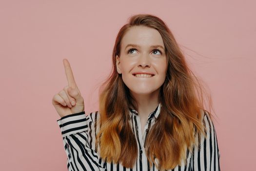 Pretty young woman in formal striped black and white blouse biting lower lip, demonstrating thinking process having pointer finger up posing against pink background. Advertisement and promotion