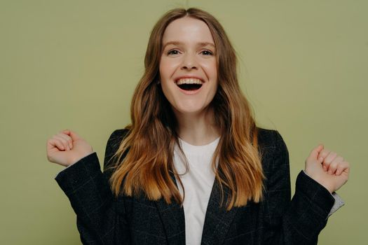 Yeah. Happy young woman with wavy brown hair holding hands up showing excitement while looking away from camera wearing dark jacket and white top, posing against light studio wall