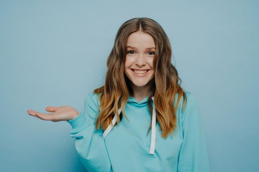 Smiling teenage girl with tipping hand shrugging shoulders standing against light blue background, cute female with wavy ombre medium length hair raising eyebrows. Human emotions concept