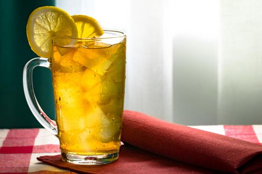 Cold tea with lemon and ice cubes in a transparent high glass cup on a red napkin