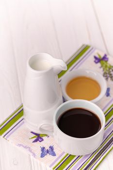 A cup of coffee, milk in a jar and honey in a bowl on a colorful napkin. White wooden background, rustic style